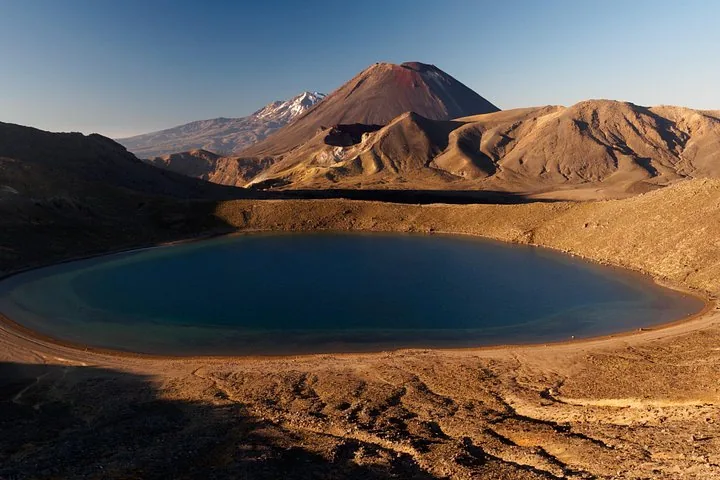 Groot blauw vulkanisch meer omringd door kaal terrein met Mount Ngauruhoe erachter.